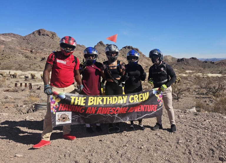 Group in helmets and adventure gear on Nevada rocks, holding 'THE BIRTHDAY CREW' banner after an epic ATV tour near Las Vegas.