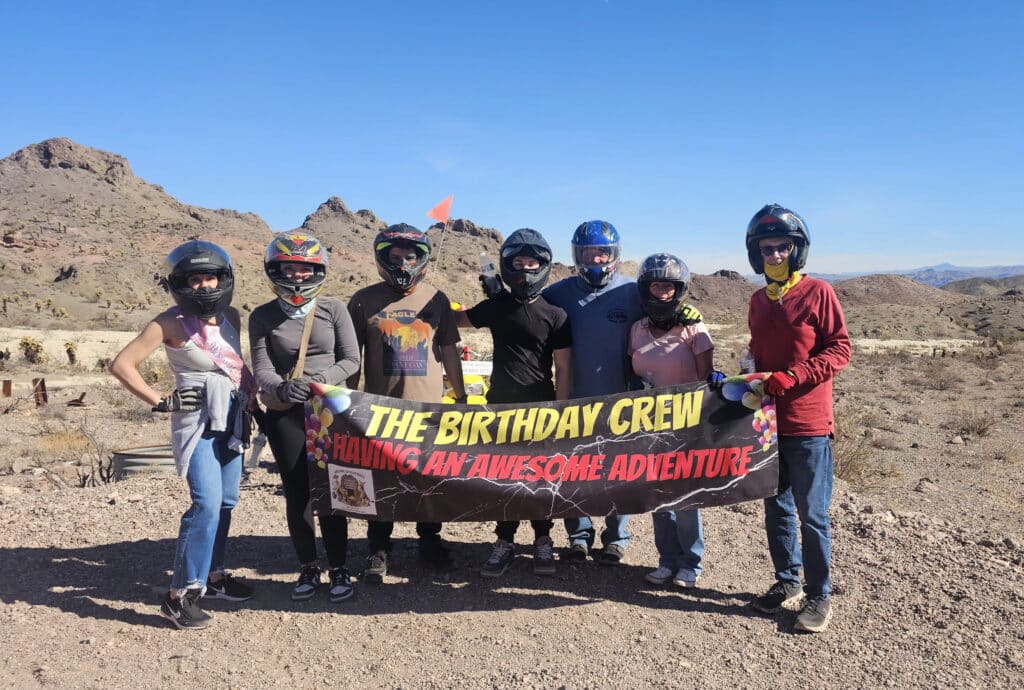 Group in helmets holds “THE BIRTHDAY CREW HAVING AN AWESOME ADVENTURE” banner on Nevada desert ATV tour near Las Vegas.