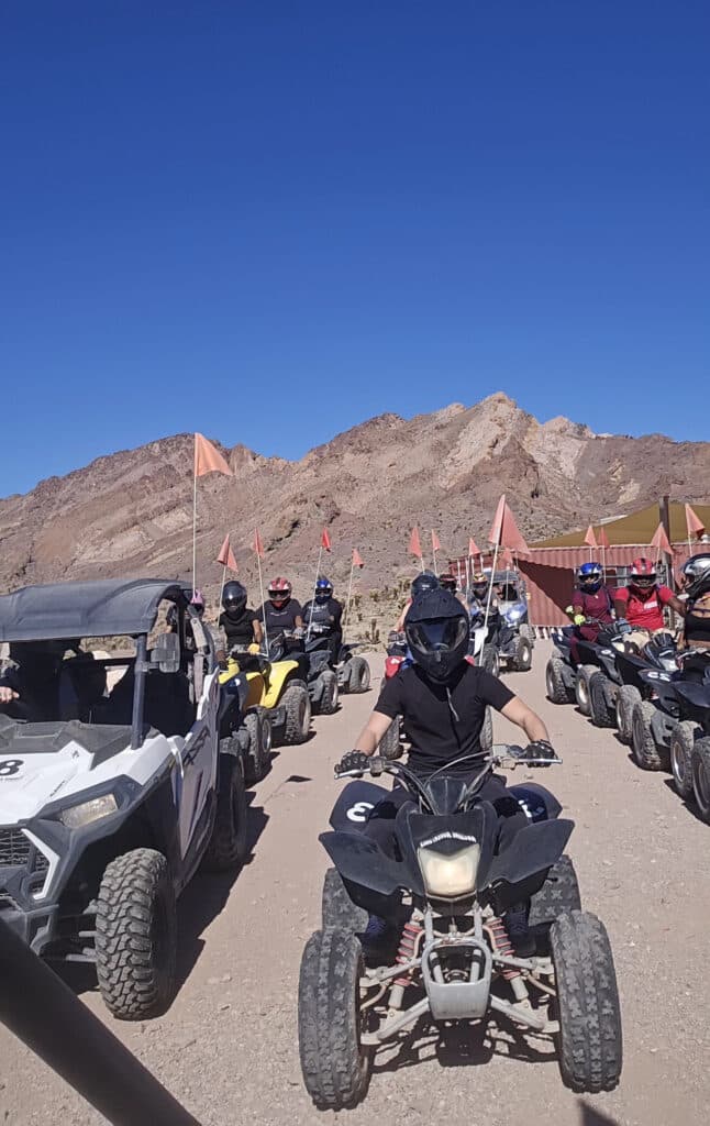 Helmeted riders enjoy a Nevada ATV tour near Las Vegas, riding off-road with orange flags and rocky mountains under a blue sky.