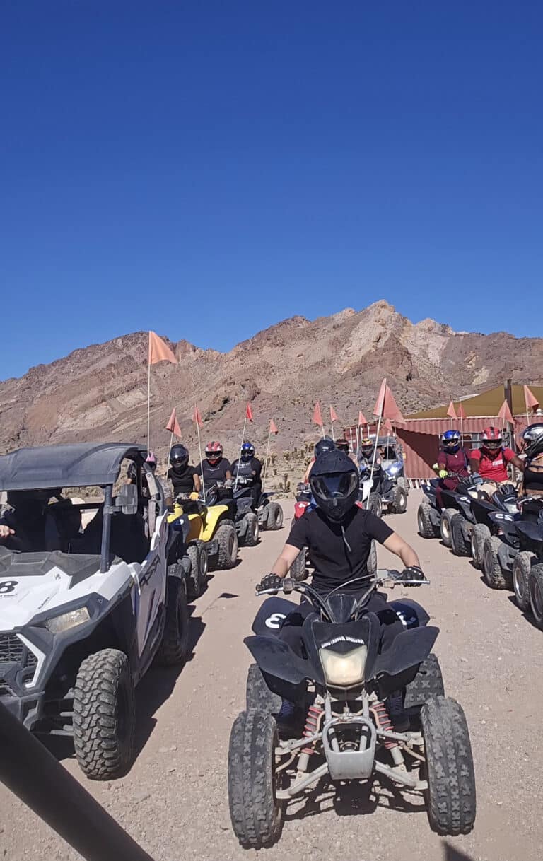 Helmeted riders enjoy a Nevada ATV tour near Las Vegas, riding off-road with orange flags and rocky mountains under a blue sky.