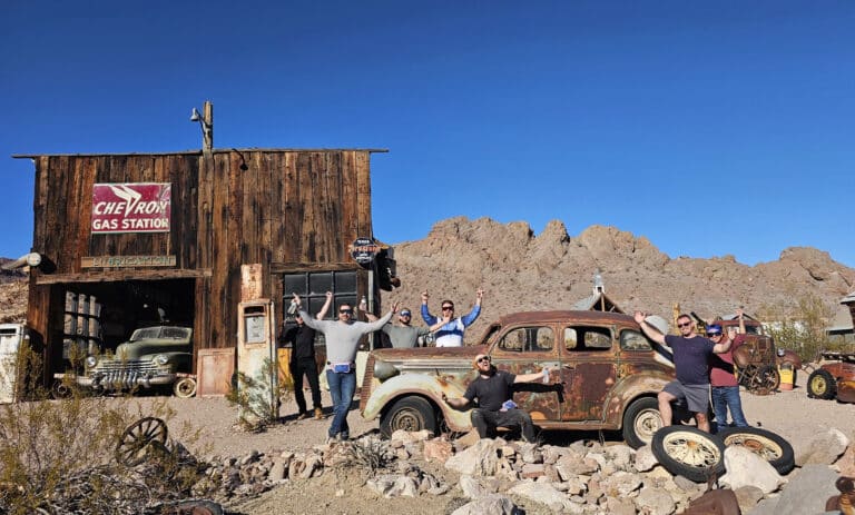 Smiling group with arms raised at a Nevada ghost town near Las Vegas, old car and gas station, perfect for ATV tours.