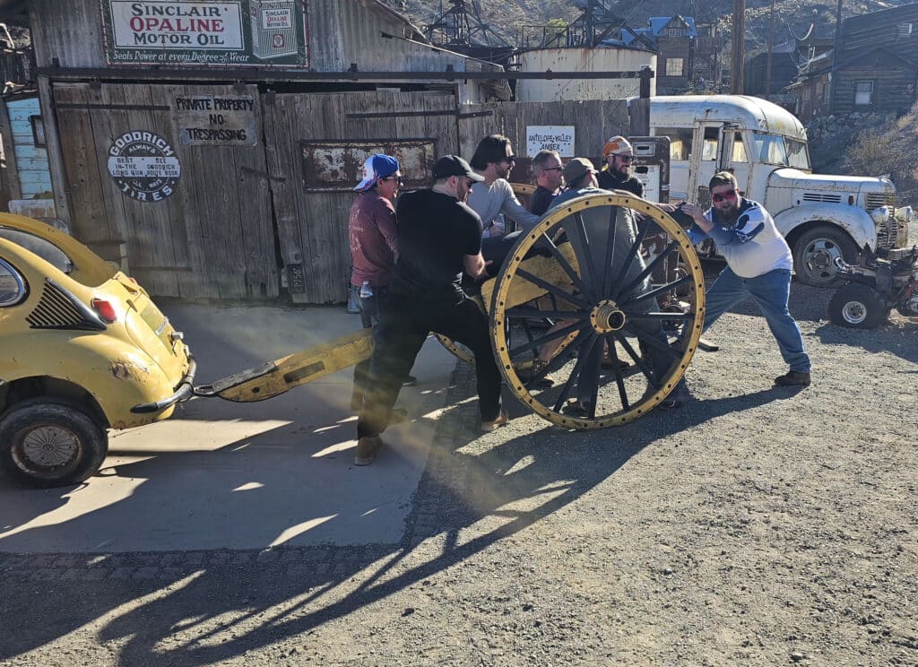 Men push a vintage wagon wheel on a yellow car near a rustic Nevada garage—Ghost Town sightseeing by Colorado River, Las Vegas.