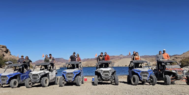 Group enjoying RZR off-road ATV tour near Nevada’s Colorado River, orange flags waving, mountains and lake under blue sky.