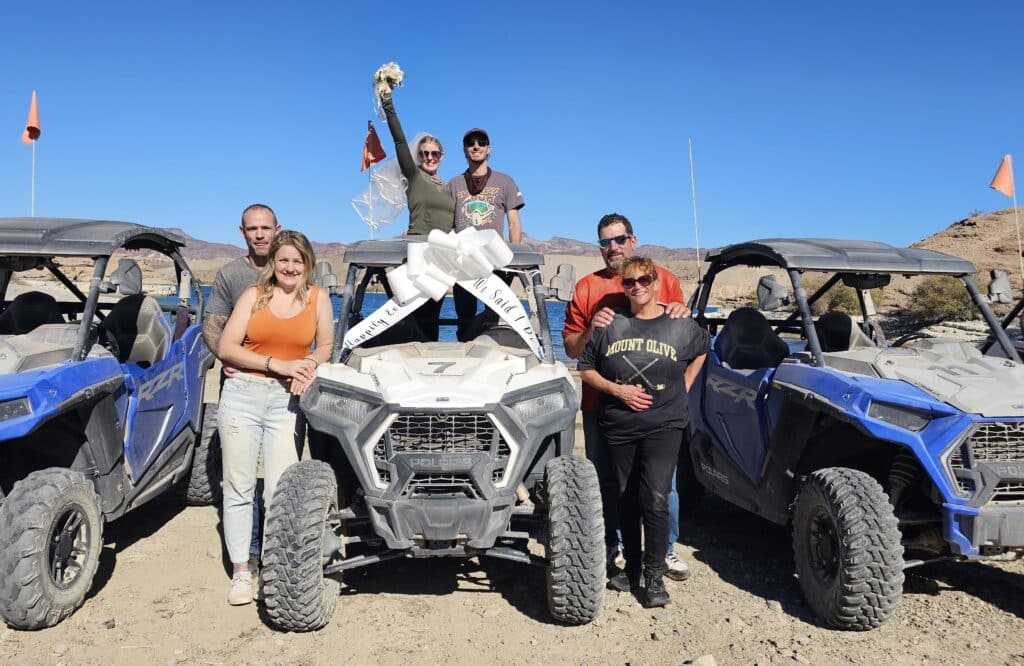 Seven friends celebrate a Las Vegas ATV tour in the Nevada desert; “Just Married” sign and bouquet on blue off-road RZR vehicles.