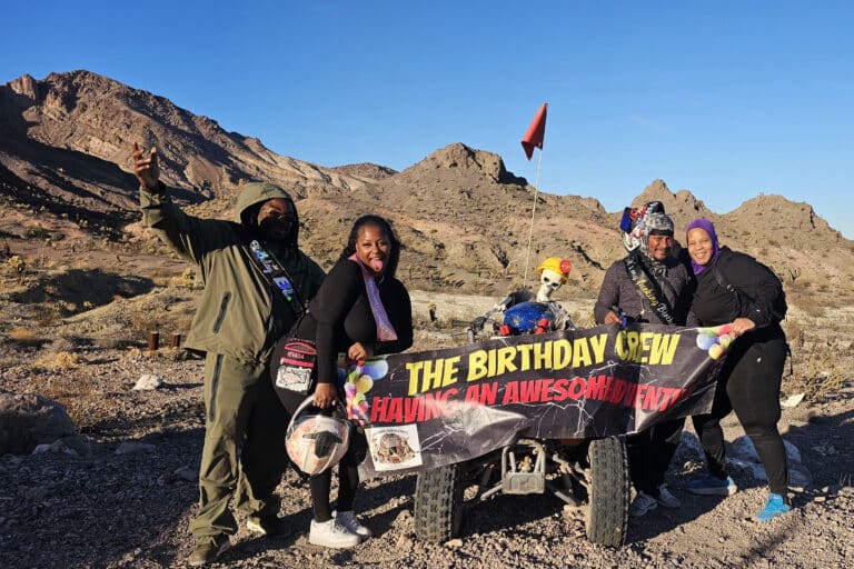 Group poses with ATV and birthday banner in Nevada desert near Las Vegas, mountains behind; perfect for ATV tours adventure.