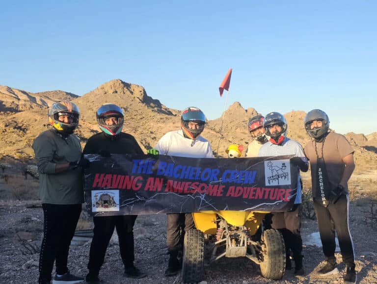 Group in helmets poses with "The Bachelor Crew" banner by rocky Nevada hills and yellow ATV on a Las Vegas RZR off-road tour.