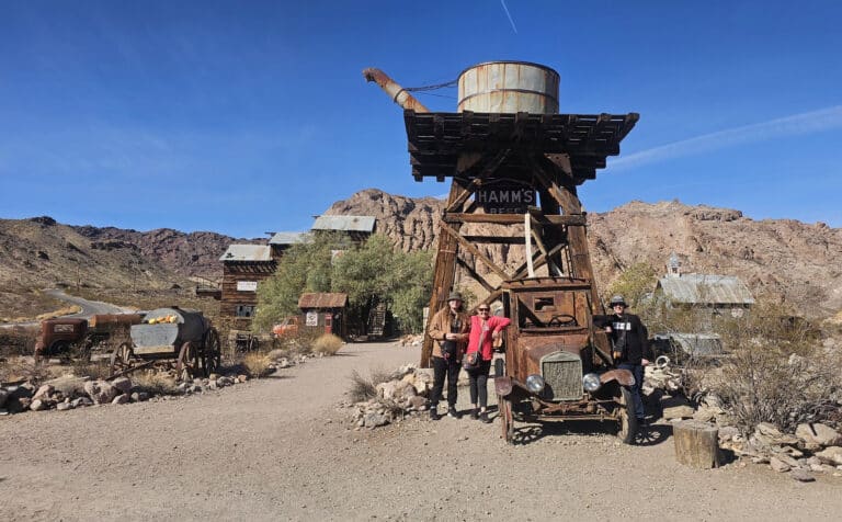 Visitors explore a Nevada ghost town near Las Vegas, standing by a rusty car and water tower on a scenic sightseeing tour.