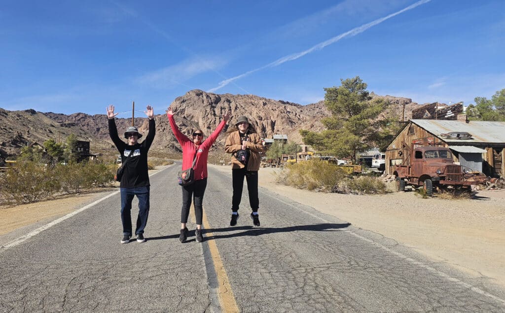 Friends pose on a Nevada desert road near old buildings, perfect for Ghost Town sightseeing and RZR off-road adventures.