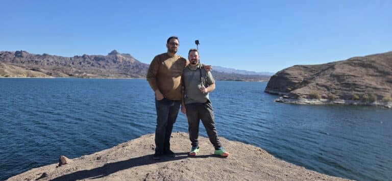 Two people pose with a selfie stick on a rocky point by the Colorado River near Las Vegas, with Nevada mountains in view.