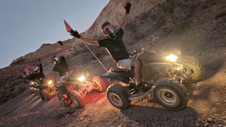 Adventurers on an ATV tour near Las Vegas ride at dusk, headlights shining, with Nevada's desert hills in the background.