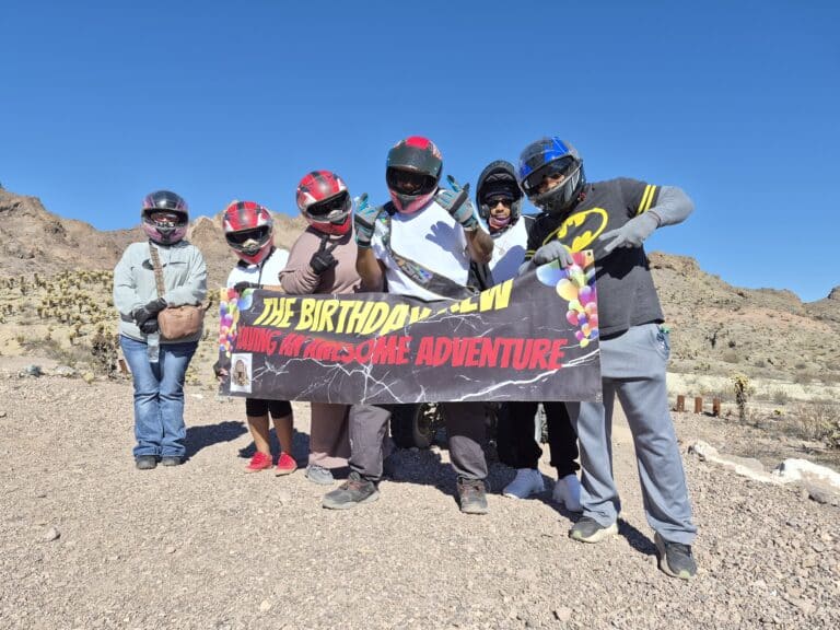 Six helmeted riders pose in the Nevada desert by Las Vegas, holding an ATV tour birthday banner with balloons and decorations.