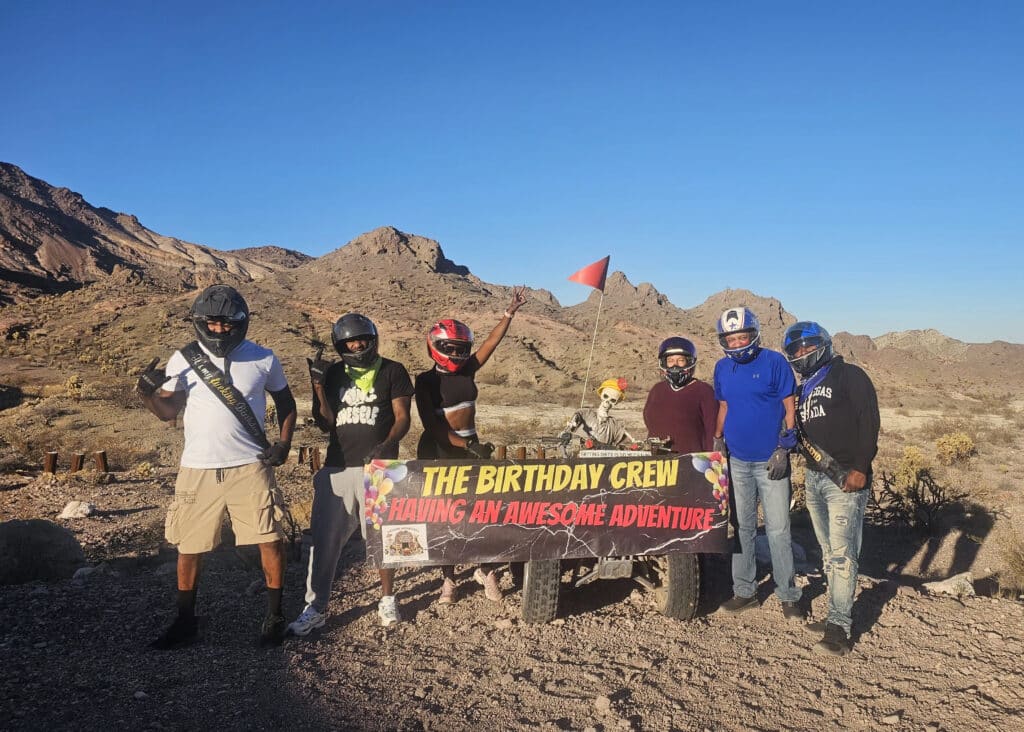 Six friends in helmets pose by an ATV and skeleton near Nevada mountains, banner reads “THE BIRTHDAY CREW,” on a Las Vegas ATV tour.