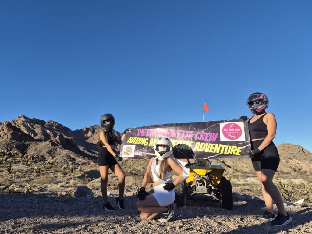 Three women on an ATV tour near Las Vegas, Nevada, pose with a fun banner; desert mountains and blue sky complete the adventure backdrop.