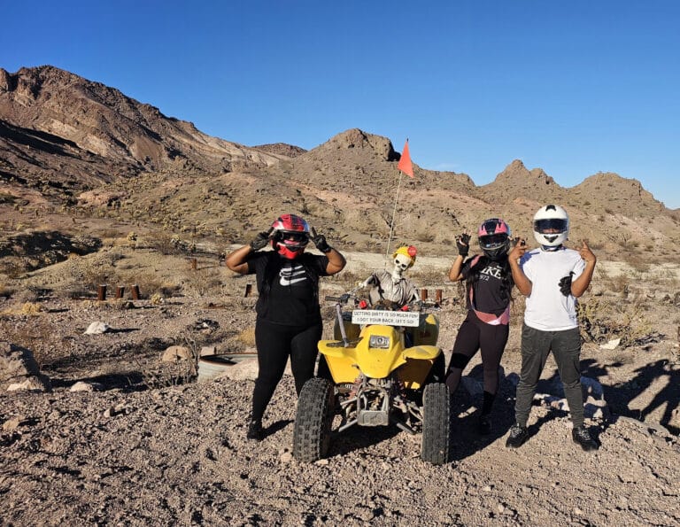 Adventurers in helmets pose by a yellow ATV on a Nevada desert tour near Las Vegas with mountains and blue sky.