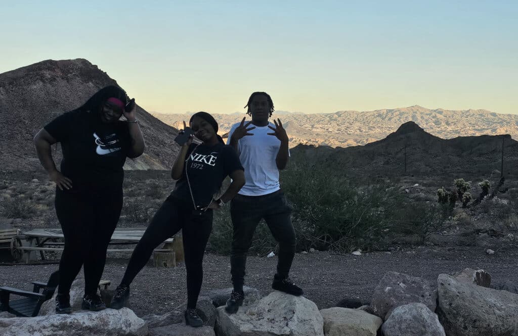 Three friends pose on Nevada desert rocks near Las Vegas mountains, dressed for a RZR off-road tour under a clear sky.