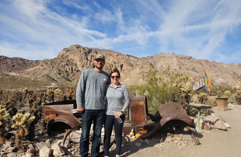 Couple by rusty vintage car parts in Nevada desert near Las Vegas, mountains, and a MOTEL sign—perfect for Ghost Town sightseeing.