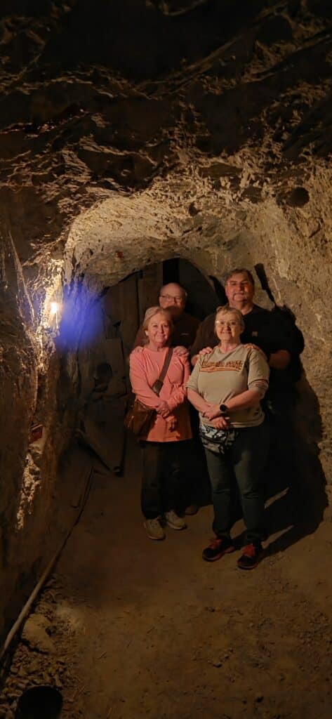 Four adults explore a dim, rocky Nevada tunnel near the Colorado River, shadows cast by a single lightbulb—perfect for Ghost Town tours.