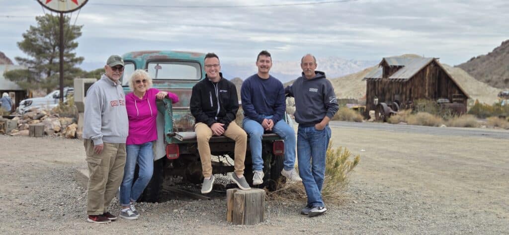 Group by an old turquoise pickup near rustic buildings, Ghost Town sightseeing in Nevada with mountains and cloudy sky.