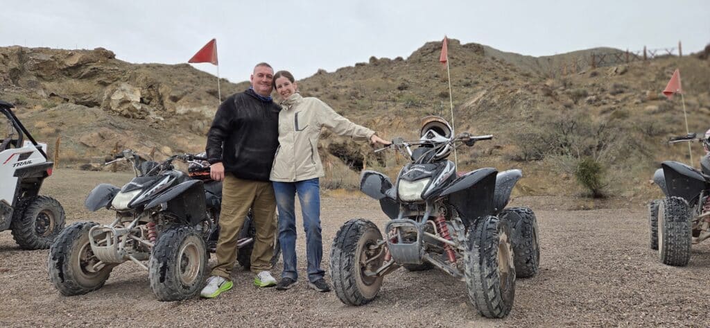 A smiling couple poses by muddy ATVs on a Nevada dirt trail, ready for Las Vegas RZR off-road and Ghost Town sightseeing adventure.