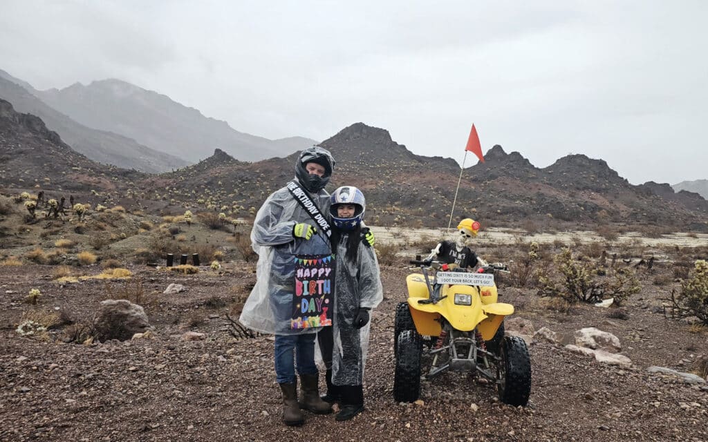 Smiling duo in rain ponchos by yellow ATV in Nevada’s desert near Las Vegas; birthday fun and off-road adventure despite the rain.