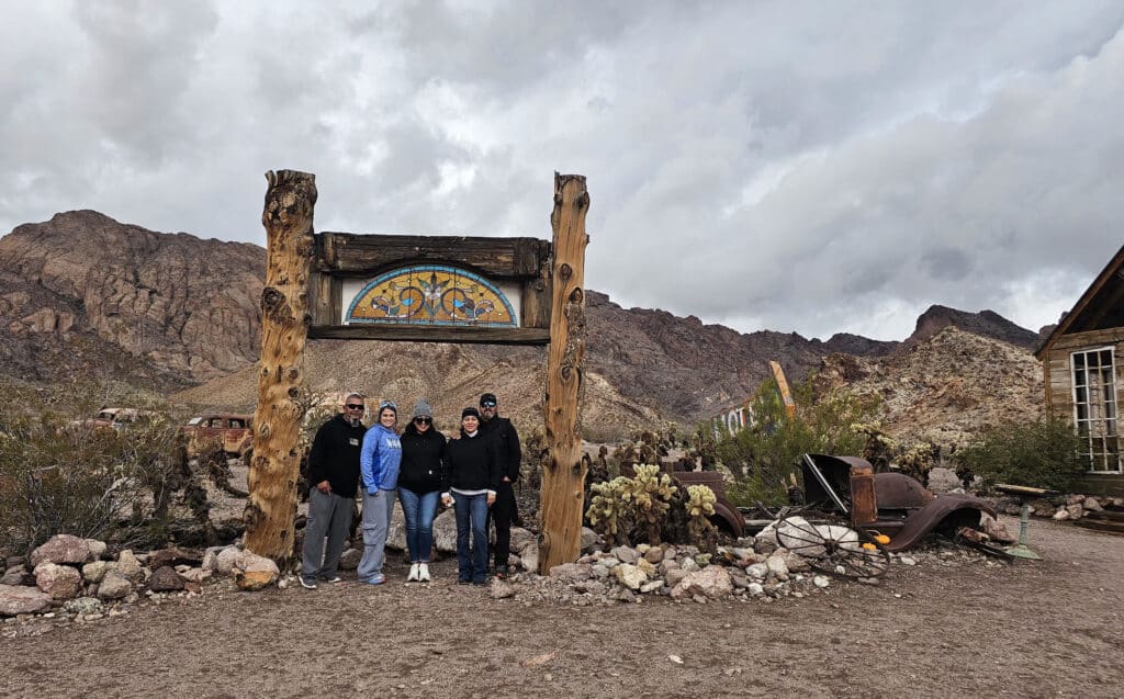 Group stands under rustic archway in Nevada desert near Las Vegas, with mountains, ghost town ruins, and old vehicle backdrop.