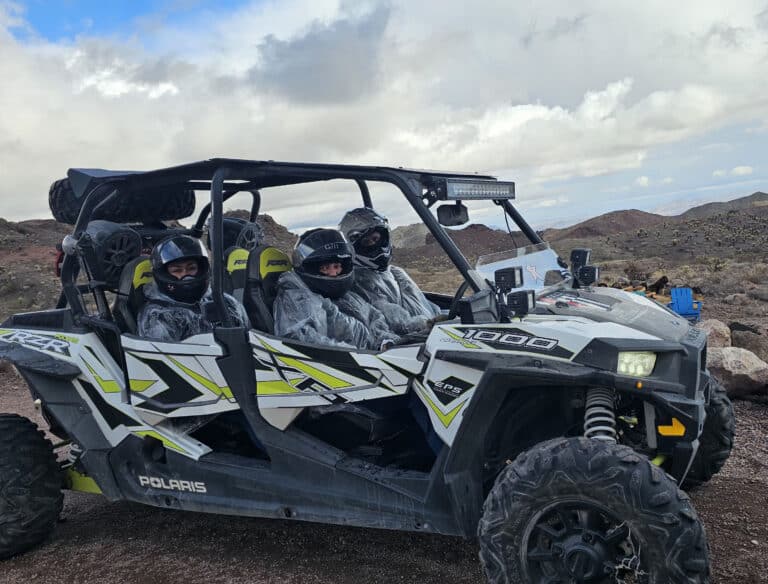 Four people in helmets enjoy a Polaris RZR ATV tour near the Colorado River, Nevada, with rocky terrain and hills under cloudy skies.