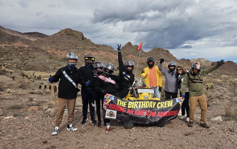 Group in helmets, outdoor gear pose in Nevada desert after ATV tour, banner reads The Birthday Crew. Smiling near Colorado River.