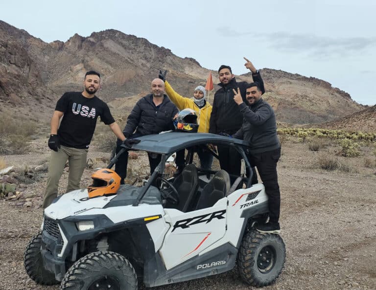 Group smiles by Polaris RZR on Nevada desert ATV tour near Las Vegas; peace signs, USA shirt, cacti, mountains in background.