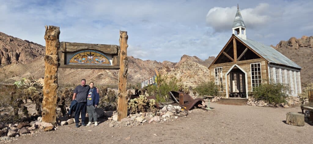 Two people at a rustic sign near a wooden chapel in a Nevada desert, ideal for Las Vegas ghost town sightseeing and ATV tours.