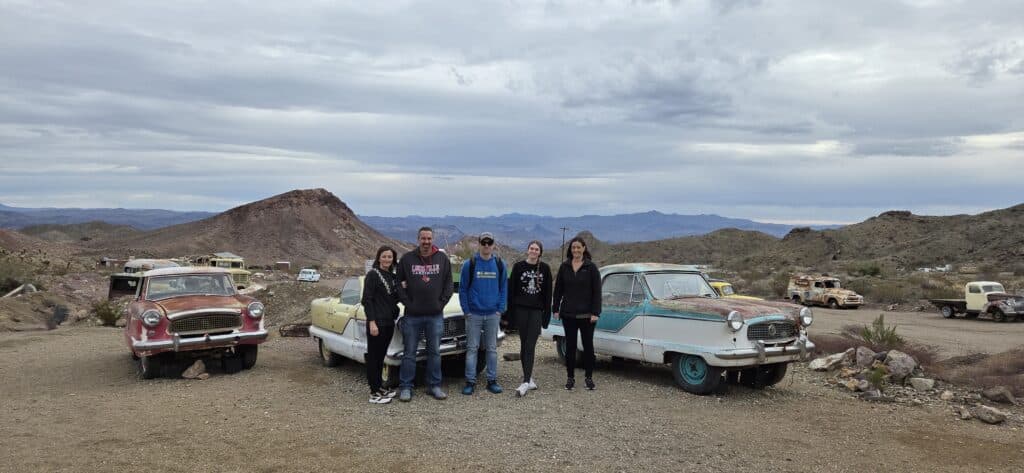 Group smiles between vintage cars in a Nevada desert near Las Vegas, with classic cars and mountains perfect for Ghost Town tours.