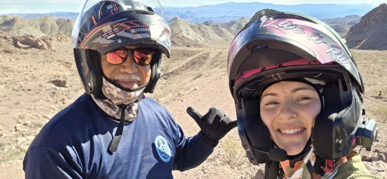 Smiling riders in helmets take a selfie on an ATV tour near Las Vegas, Nevada desert with mountain views under blue sky.