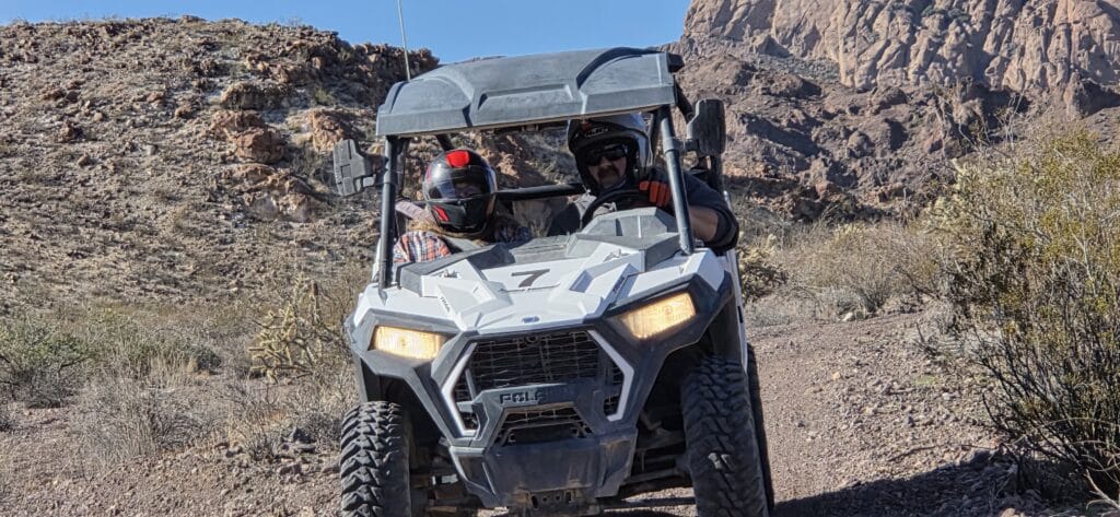 Riders in helmets on a white ATV explore a rocky Nevada desert trail near Las Vegas with mountain views under blue sky.
