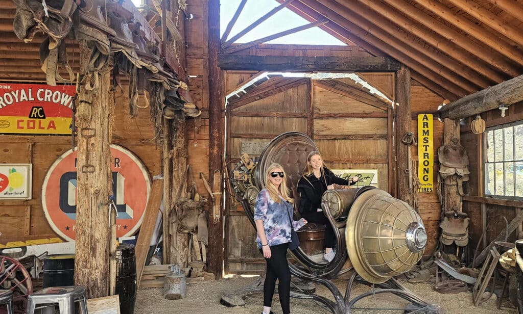 Two women smile in a Nevada ghost town barn with vintage western decor and a quirky sculpture, perfect for Las Vegas sightseeing.