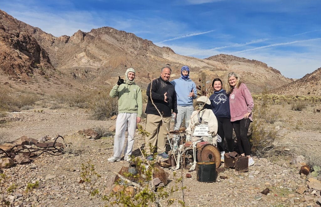 Group smiles by skeleton drummer statue in Nevada desert near Las Vegas, with mountains—perfect for Ghost Town sightseeing tours.