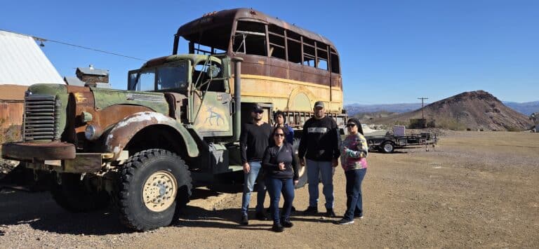 Group poses by a vintage rusted double-decker bus in Nevada desert near mountains, ideal for Ghost Town sightseeing tours.