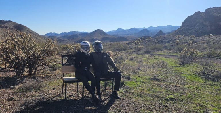 Two riders in motorcycle gear relax on a desert bench near cacti and mountains in Nevada, close to Las Vegas ATV tour trails.