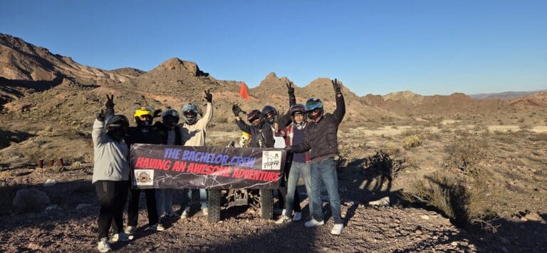 Group in helmets and jackets holds “The Bachelor Crew Having An Awesome Adventure” banner on Nevada desert ATV tour near Las Vegas.