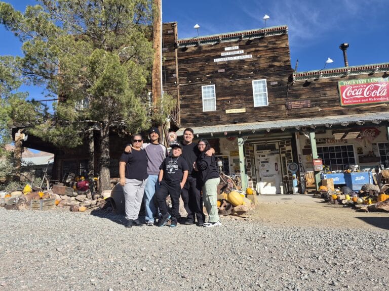 Group smiles by rustic Nevada ghost town with vintage signs, pumpkins; great spot for Las Vegas ATV tours and Colorado River views.