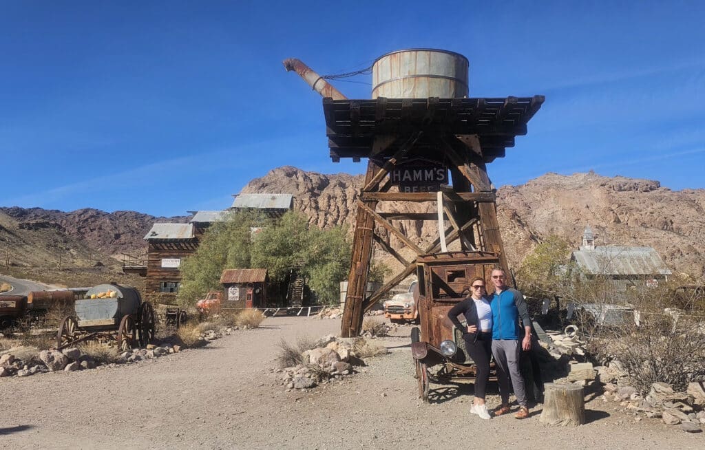 Ghost Town sightseeing near Las Vegas: two visitors under HAMMS BEER water tower, vintage wagons, and rocky Nevada hills.