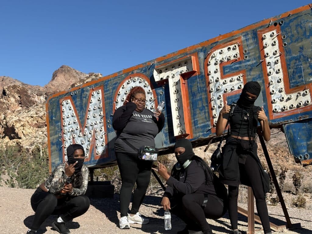 Four women in black outfits pose by a weathered motel sign near Nevada’s rocky desert, perfect for Las Vegas ghost town tours.