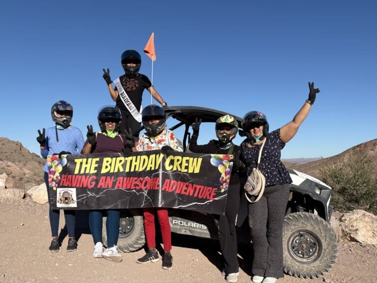 Group of six in helmets pose by RZR in Nevada desert on a Las Vegas ATV tour, holding "The Birthday Crew" banner and smiling.