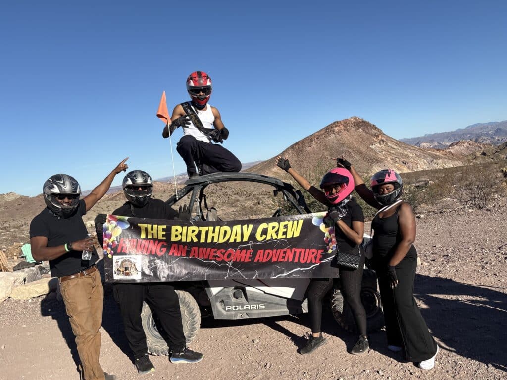 Group in helmets and outdoor gear poses with banner by ATV during a Nevada RZR off-road adventure near Las Vegas desert.
