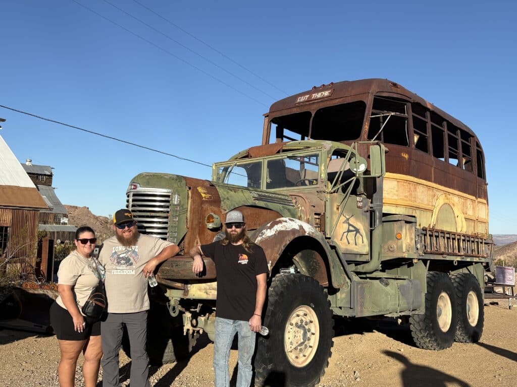 Three adults pose smiling by a rusted military-style truck in Nevada desert, perfect for Ghost Town sightseeing near Las Vegas.