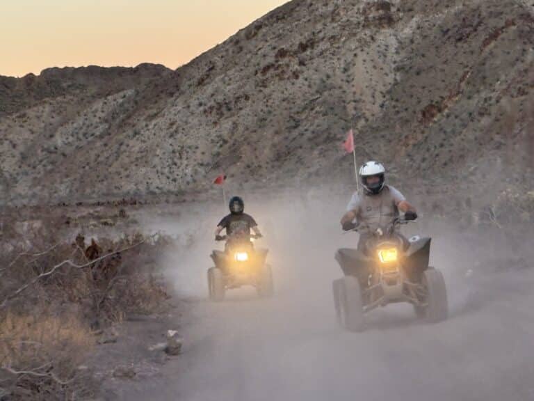 ATV riders explore a dusty Nevada trail at sunset near rugged hills, headlights on, red flags flying—perfect for Las Vegas ATV tours.