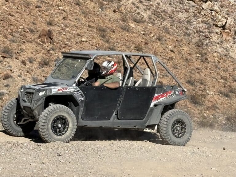 Helmeted rider drives an ATV near a rocky Nevada hillside, kicking up dust on a Las Vegas RZR off-road tour trail.