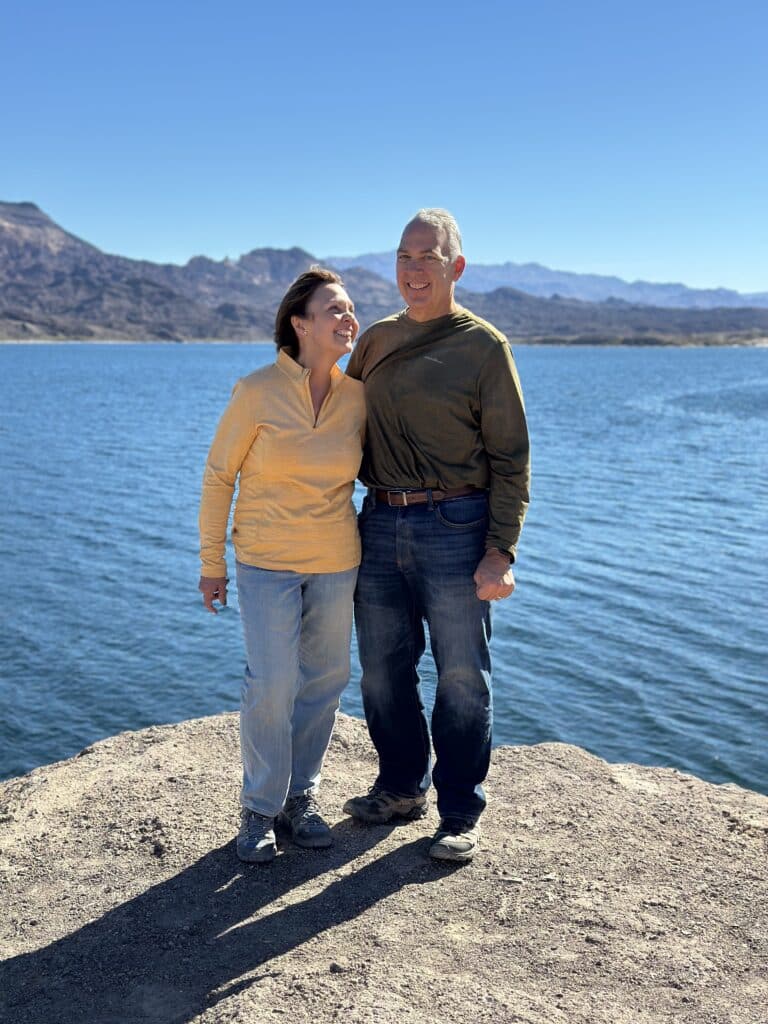 Smiling couple on Nevada’s Colorado River shore near Las Vegas, mountains behind. Yellow & green sweaters, clear sky.
