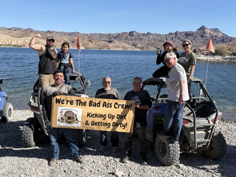 Smiling group by ATVs on the Colorado River near Las Vegas, holding sign after a Nevada RZR off-road tour.