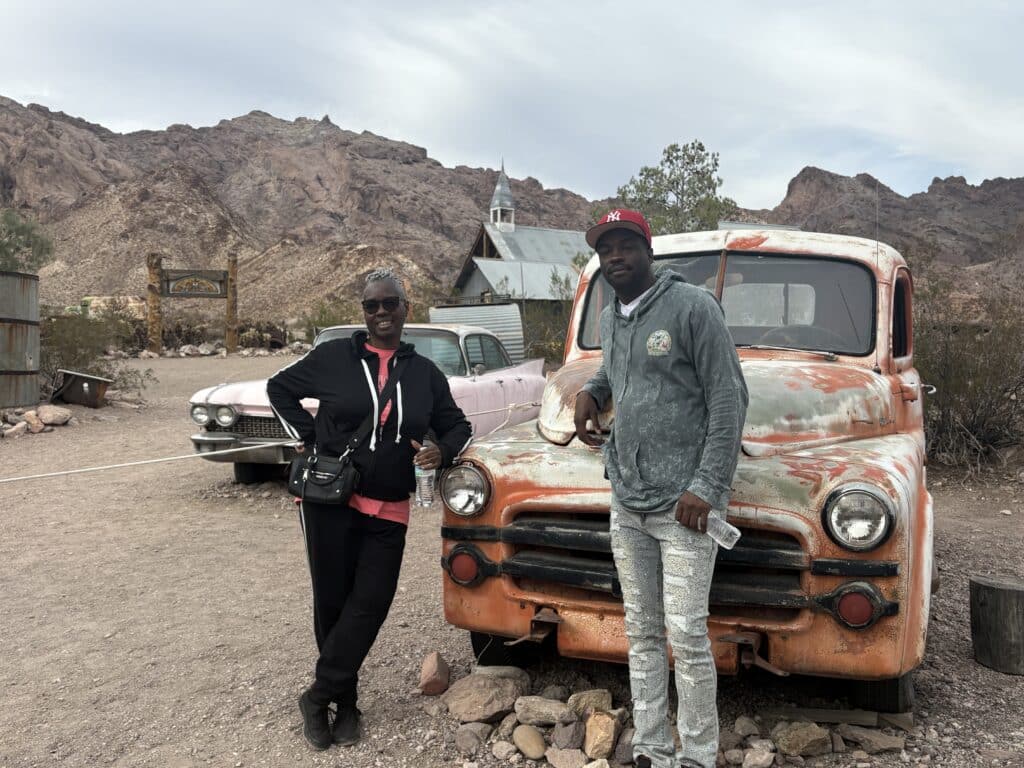 Two people smile by a rusty orange truck in a Nevada desert ghost town near Las Vegas, with old cars and scenic mountains behind.