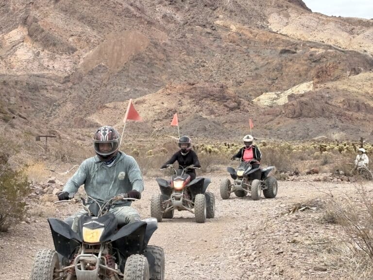 Adventurers on ATVs with red flags ride a rocky Nevada trail near Las Vegas, helmets on, exploring desert terrain and arid mountains.