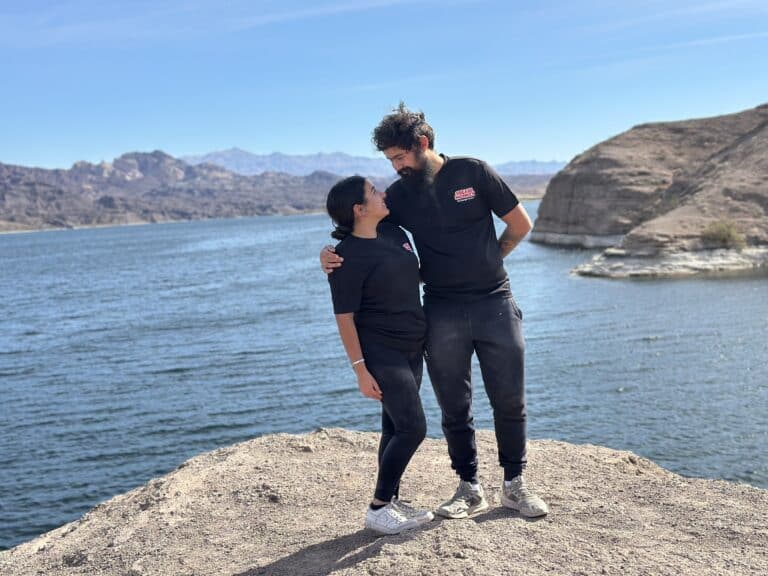 Two people in black outfits smile on rocky ledge above Colorado River near Las Vegas, Nevada with blue sky and mountain views.